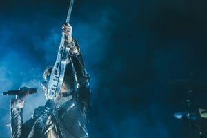 man holding electric guitar and microphone in front of lights inside dim room with smoke